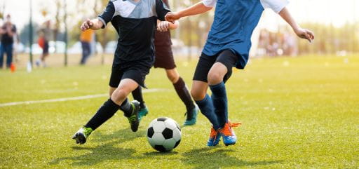 Photo of children playing soccer on a grass field outside. The image is just of the players legs and the ball. 
